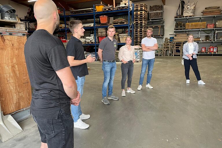 Junge Leute stehen im Halbkreis in einer Halle mit Holz und Metall und Unterhalten sich