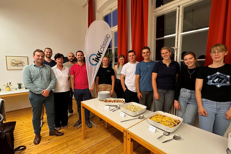 Eine Gruppe mit mehreren Menschen, die sich in einer Reihe aufgestellt haben und vor einem Tisch mit frisch gekochtem Essen stehen