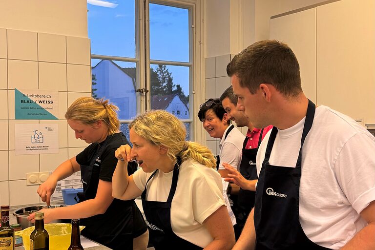 Drei Personen stehen vor einem Herd mit Töpfen und Pfannen. Sie Kochen zusammen und probieren von dem Essen