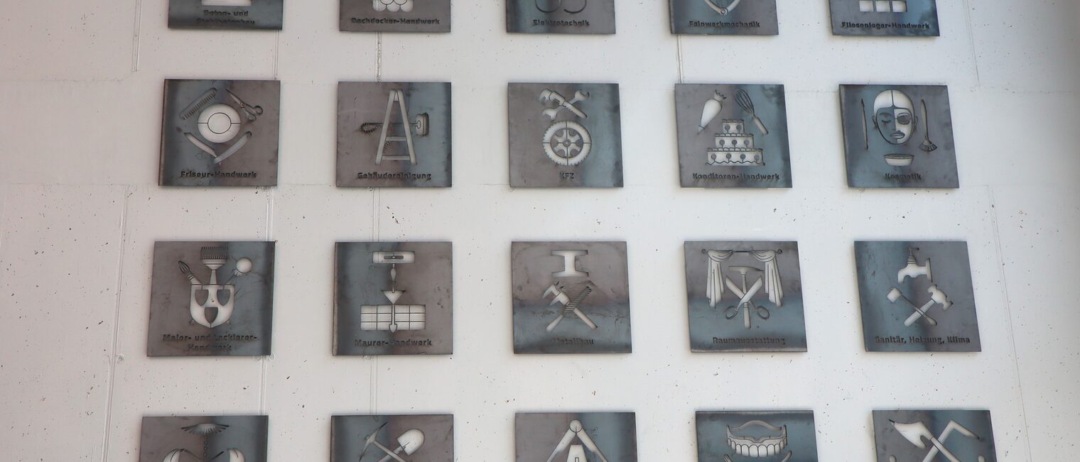 20 verschiedene Zunftzeichen des Handwerks werden gezeigt. Sie hängen in vier Reihen an der Wand. 