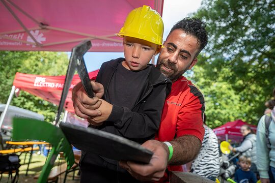 Kind mit Werkzeug bearbeitet mit Hilfe eines Handwerkers eine Schiefernplatte.