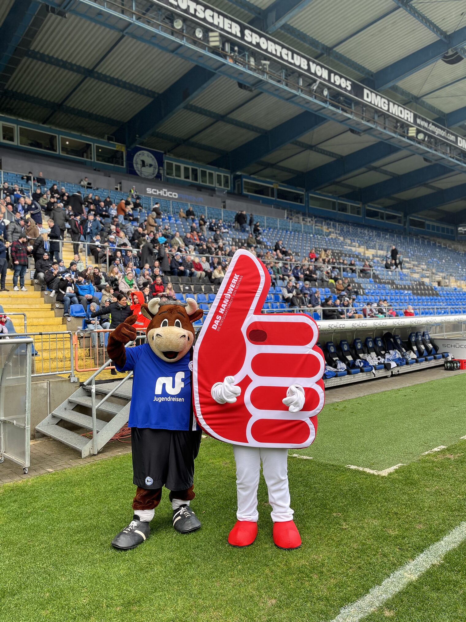 Lohmann (Maskottchen Arminia) und Daumenik (Handwerksmaskottchen) stehen zusammen in der Schüco-Arena vor der Tribüne.