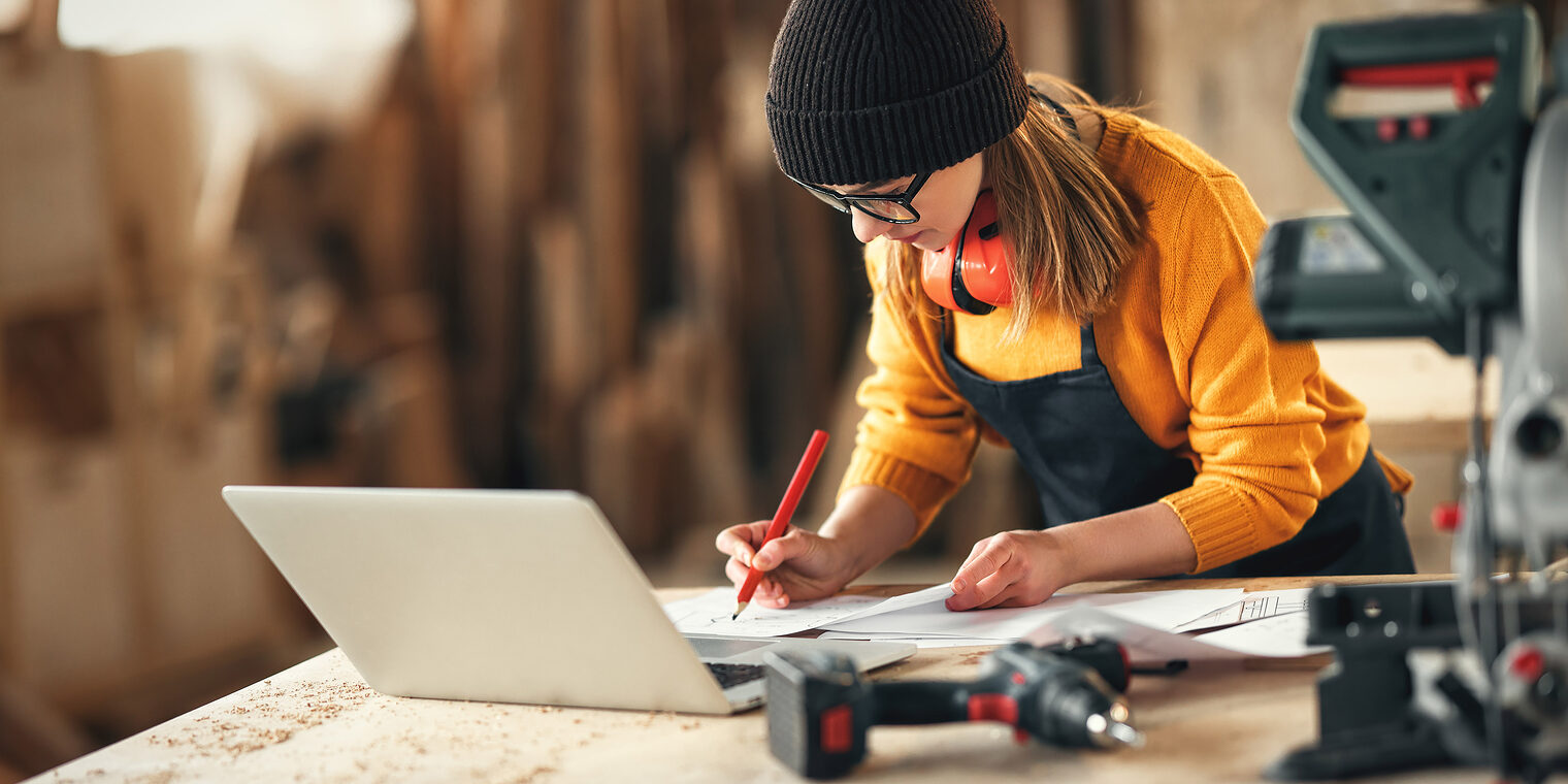 Focused young skilled craftswoman making notes in papers while standing at table with laptop and carpentry instruments in workshop Schlagwort(e): craftswoman, carpenter, joiner, work, paper, take note, laptop, workshop, artisan, craft, woodworker, woman, carpentry, small business, check, write, document, professional, female, occupation, self employed, job, skill, project, device, craftsmanship, lifestyle, woodwork, manufacture, plan, draft, entrepreneur, joinery, handicraft, master, profession, busy, paperwork, maker, labor, worker, indoors, blur, selective focus, girl, young